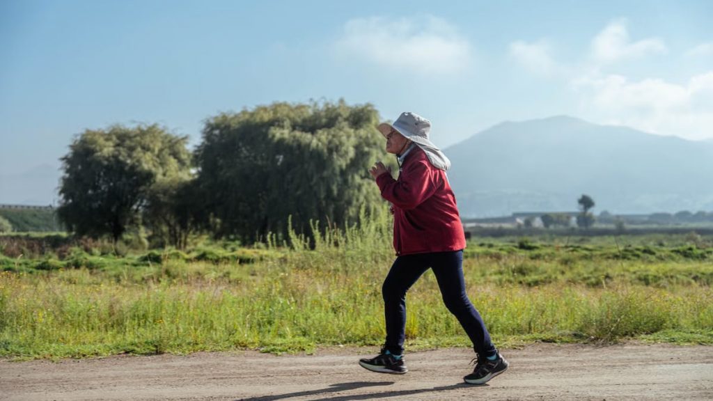 A woman in sportswear jogs along a rural road, surrounded by lush countryside and mountains.